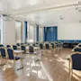 Konferencelokale med rækker af blå stole og borde, vendt mod en stor projekterskærm. Stærkt sollys filtrerer gennem høje vinduer med blå gardiner, og oplyser trægulvet.