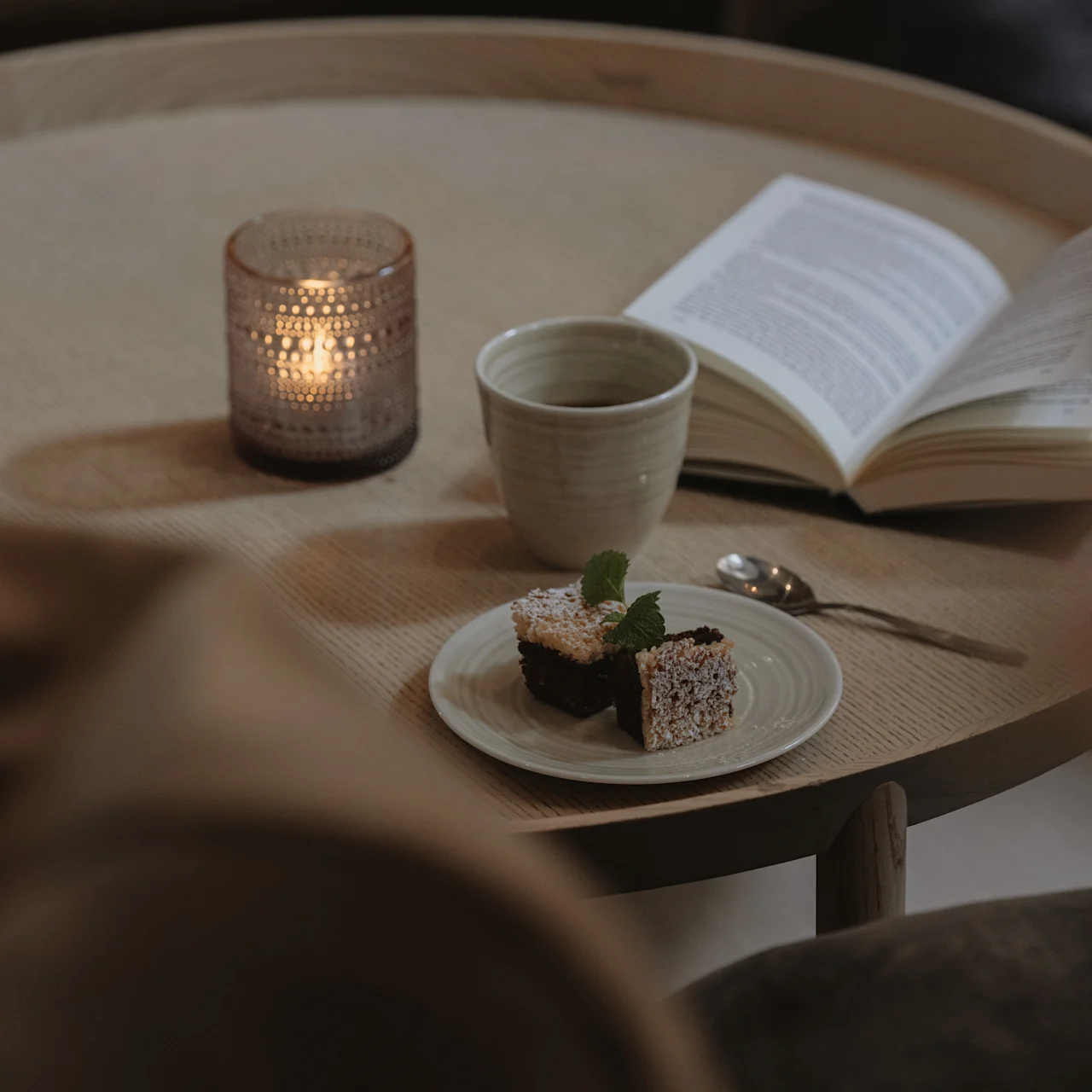 Ett ljus lyser på ett runt träbord bredvid en öppen bok, en kopp och en desserttallrik, vilket skapar en mysig atmosfär.