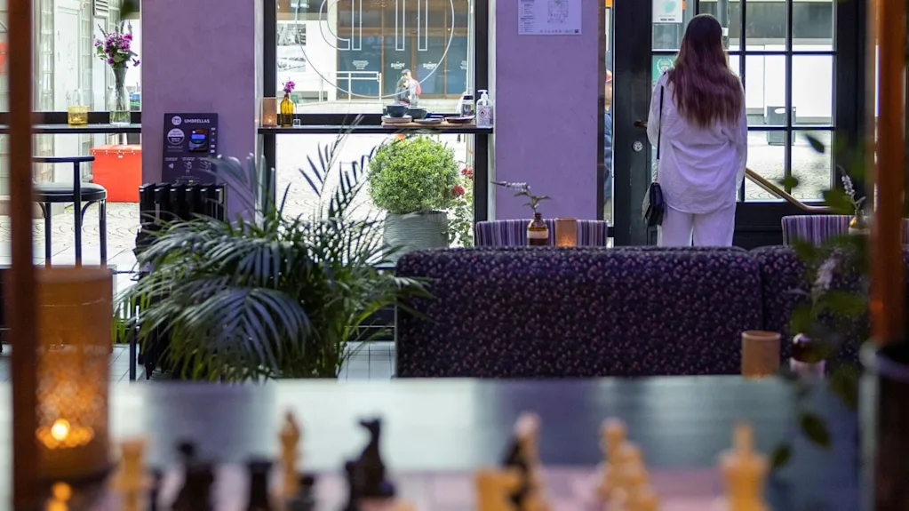 A chessboard sits idle on a table in a cozy cafe, while a person in white stands near the entrance. Plants and soft lighting add to the ambiance. Text on umbrella dispenser: "UMBRELLAS".