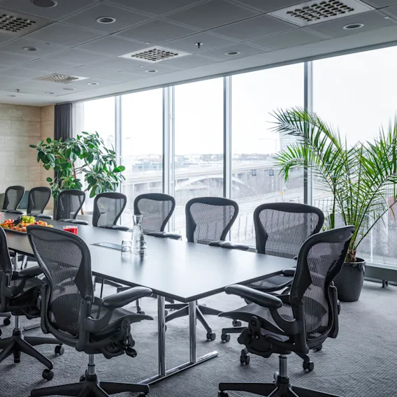 A long conference table is surrounded by ergonomic chairs, with fruit platters and drinks on top. Large windows and plants create a bright, modern office environment.