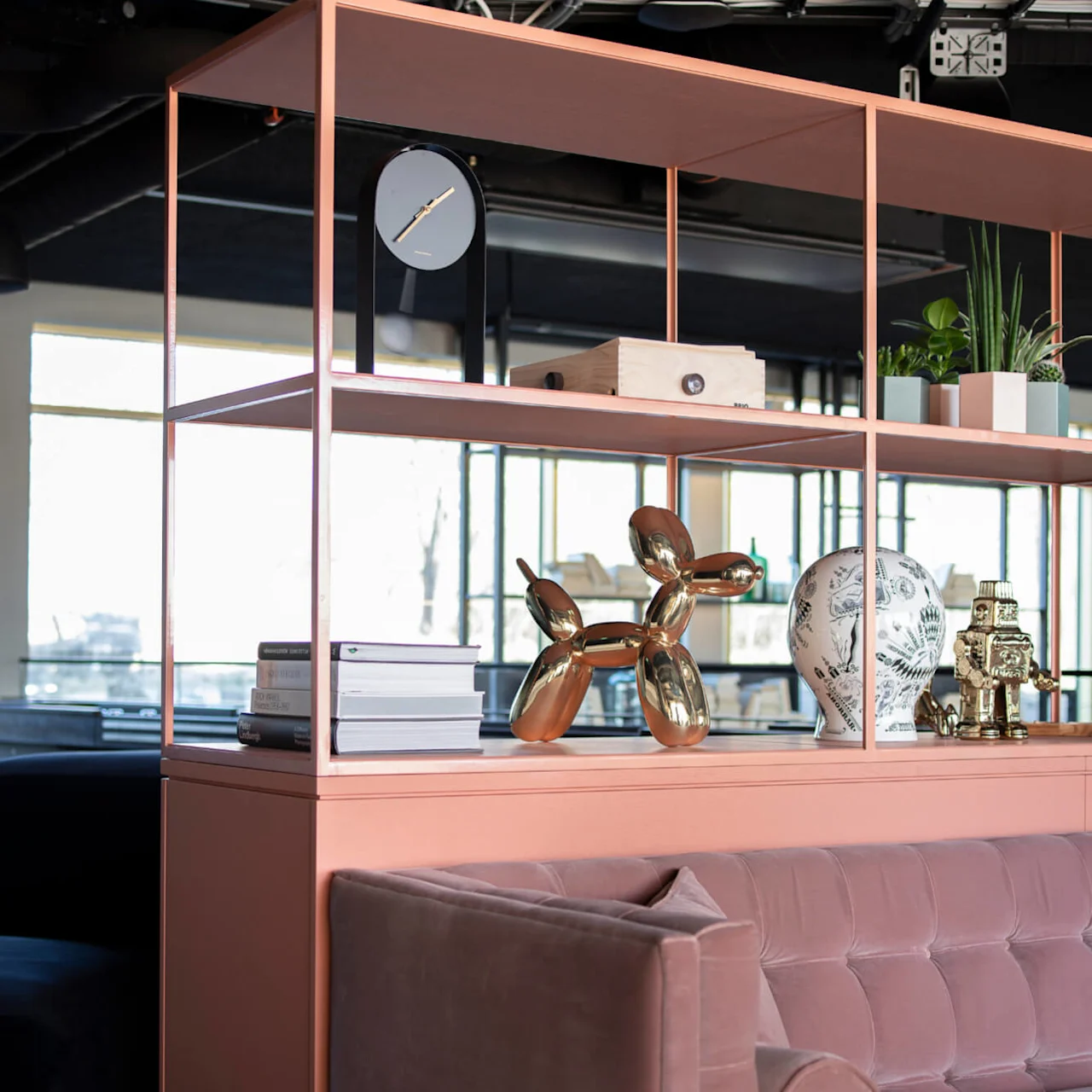 Bookshelf displaying a modern clock, shiny balloon dog sculpture, decorative white sphere, and plants. Soft pink cushioned sofa sits below. Bright daylight filters through large windows.