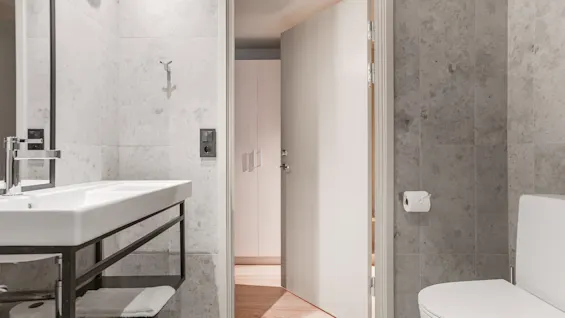 A modern bathroom with a white sink, a toilet, and an open door leading to a room with a closet. The walls are tiled.