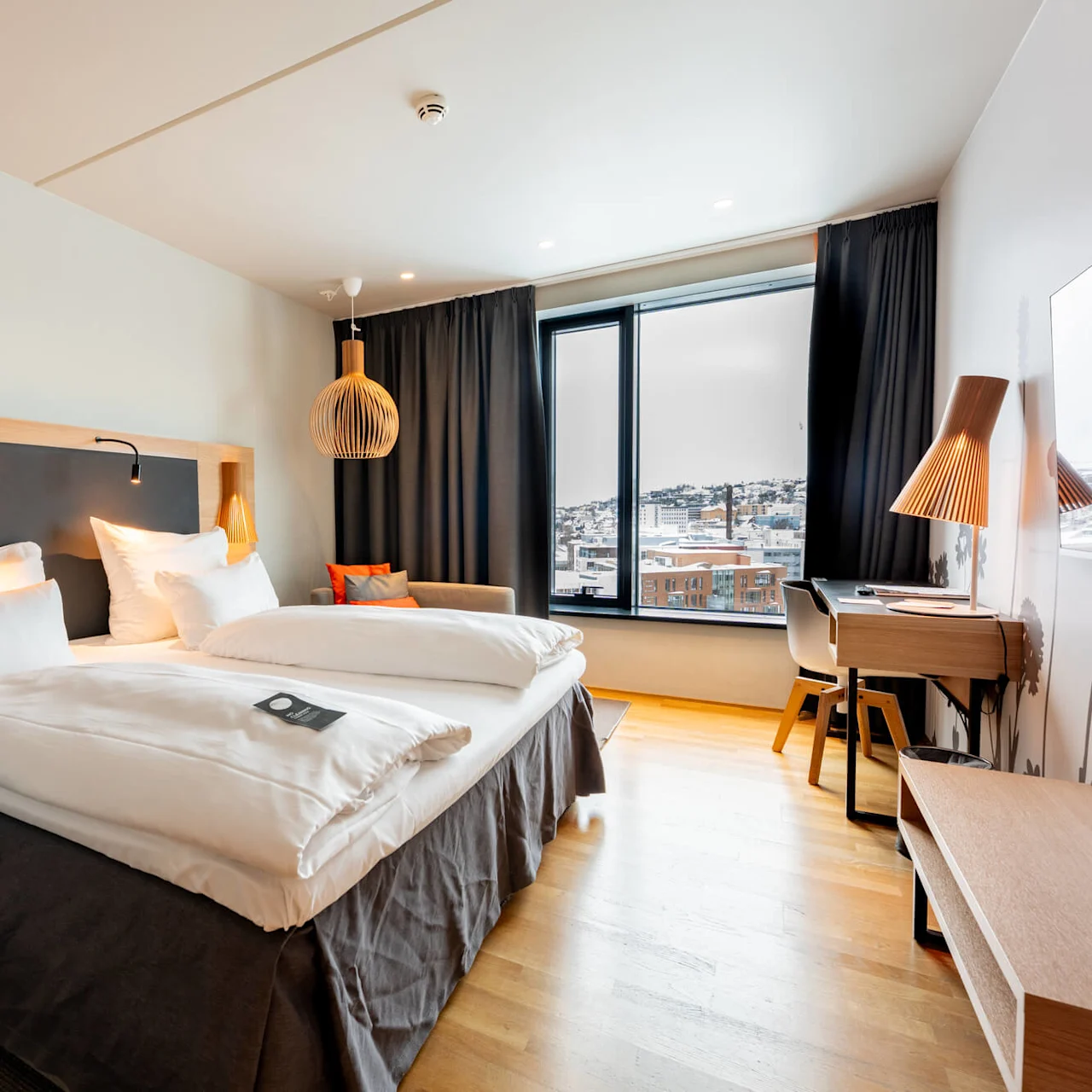 En pænt redt seng med hvidt sengetøj står i et moderne hotelværelse. Et vindue viser et sneklædt bylandskab, suppleret med stilfulde møbler i træ og blød belysning. Ingen tekst til stede.