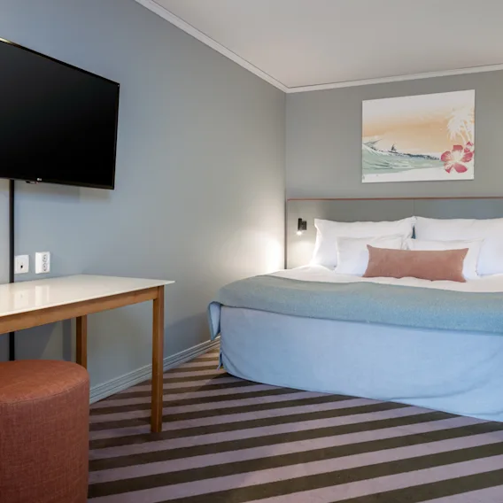 En seng med hvitt og blått sengetøy er plassert i et hotellrom. Et maleri henger over sengen. Et bord og en krakk står overfor, med en flatskjerm-TV på veggen.