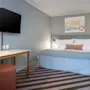 En seng med hvitt og blått sengetøy er plassert i et hotellrom. Et maleri henger over sengen. Et bord og en krakk står overfor, med en flatskjerm-TV på veggen.