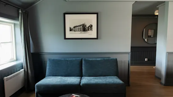 Blå sofa står i en stue, overfor et vindue med lukkede gardiner, under et indrammet sort-hvidt fotografi. I nærheden står et lille bord med et par bøger.