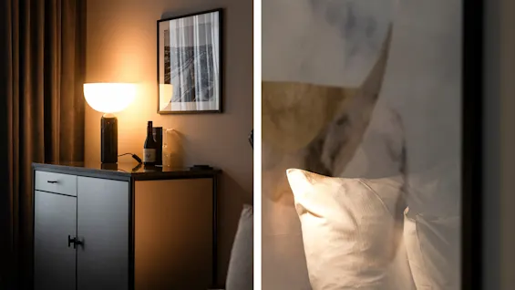 Et sidebord med en lysende lampe, flaske og vinglas står i et svagt opplyst rom med gardiner og en indrammet billede. En pude ses i en refleksion.