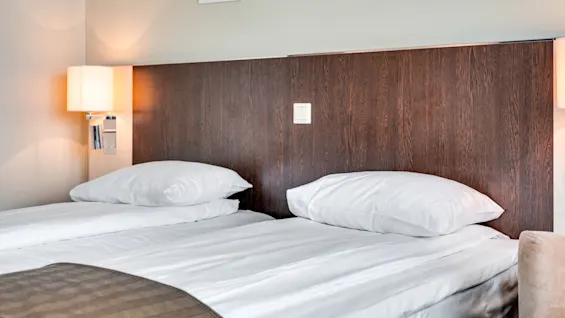 A neatly made bed with white linens and two pillows against a wooden headboard, illuminated by a bedside lamp, in a clean, modern bedroom.
