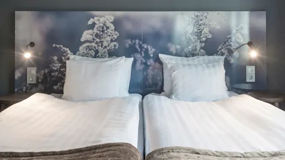 Två prydligt bäddade enkelsängar har vita sängkläder under en huvudgavel med blommigt motiv. Väggmonterade lampor belyser båda sidor, kompletterade av ett mörkt, minimalistiskt rum.