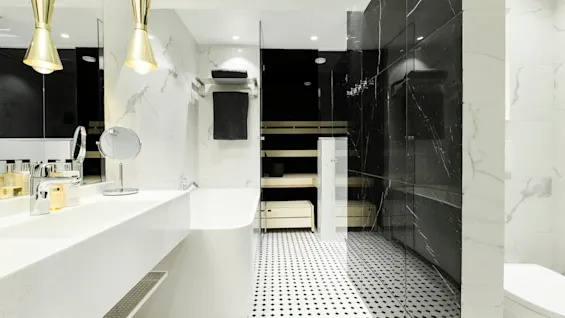 White bathtub and sink stand in a modern bathroom, accented by gold fixtures. Behind glass, a black-marble shower area contrasts with the patterned tile floor. Towels hang on the wall.