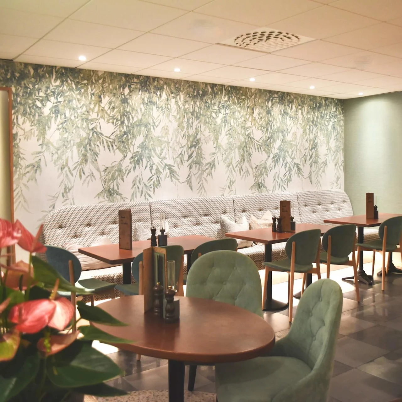 Bord och stolar fyller en mysig restaurang, dekorerad med blommig tapet. Mjuk belysning skapar en varm atmosfär. Rosa blommor pryder ett bord och förhöjer den eleganta miljön.