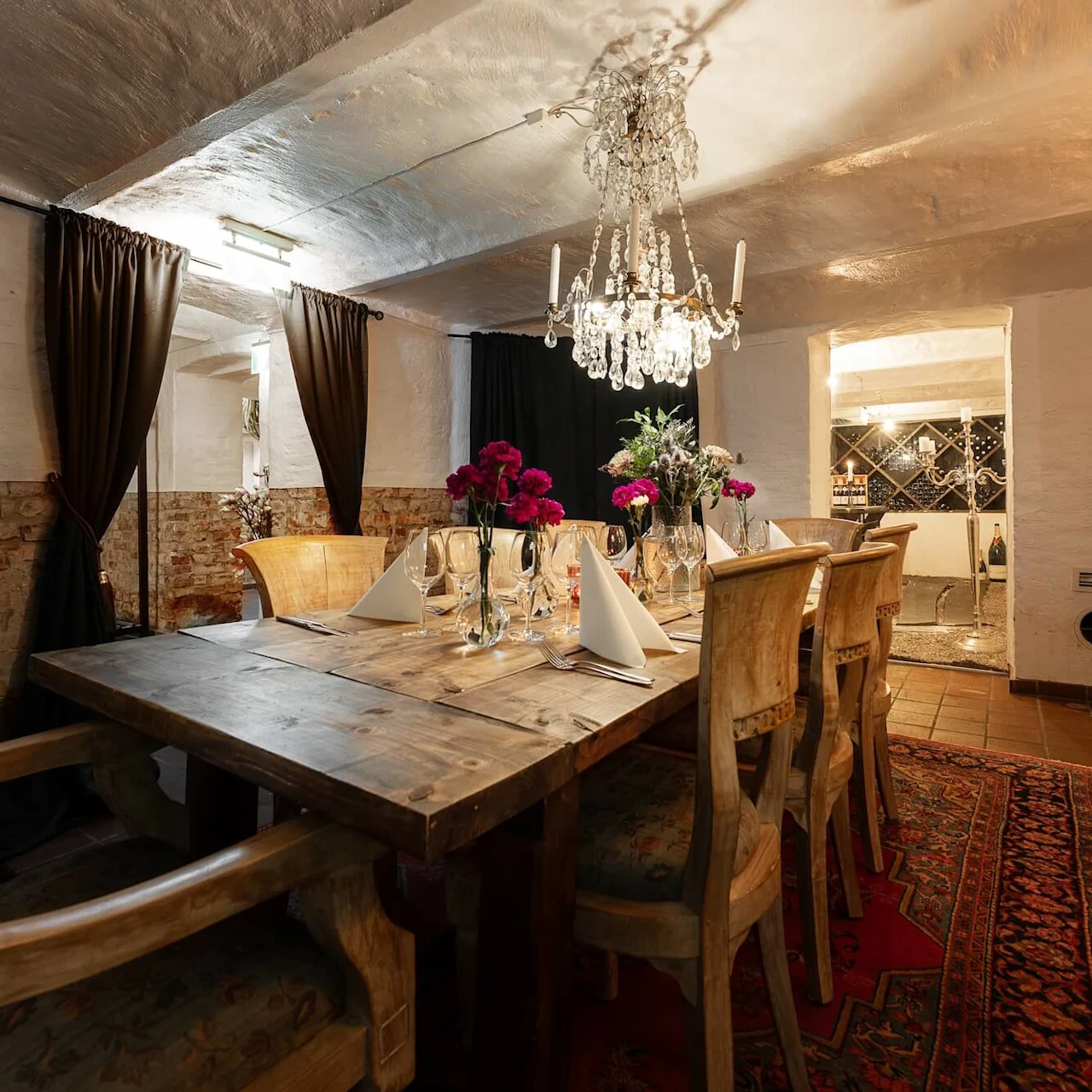 A wooden dining table is set with glasses, napkins, and flowers under a chandelier. It is in a cozy, dimly lit room with brick walls and heavy curtains.