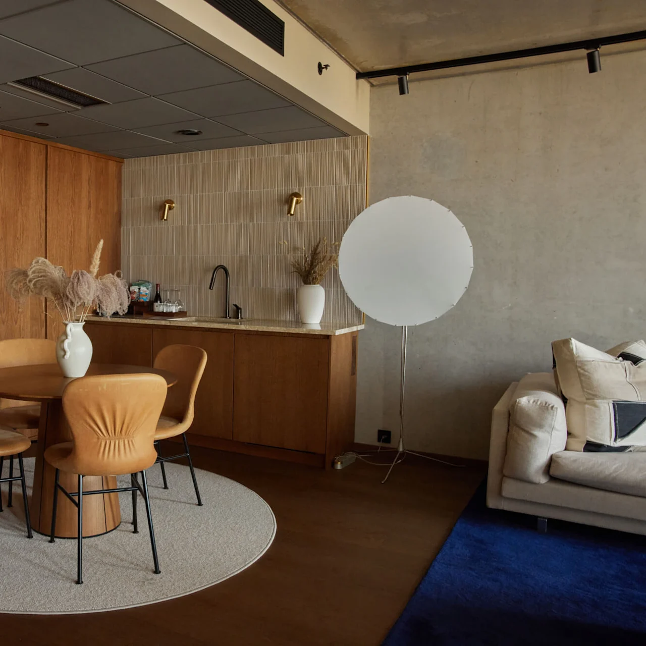 Ett runt träbord med fyra stolar står på en rund matta bredvid en pentry. En stor, vit cirkelformad lampa står nära en beige soffa.