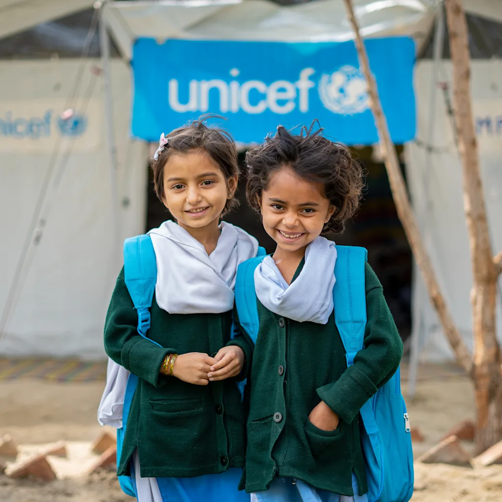 To smilende barn med ryggsekker står foran et telt med teksten unicef og MPBAGLPUR.