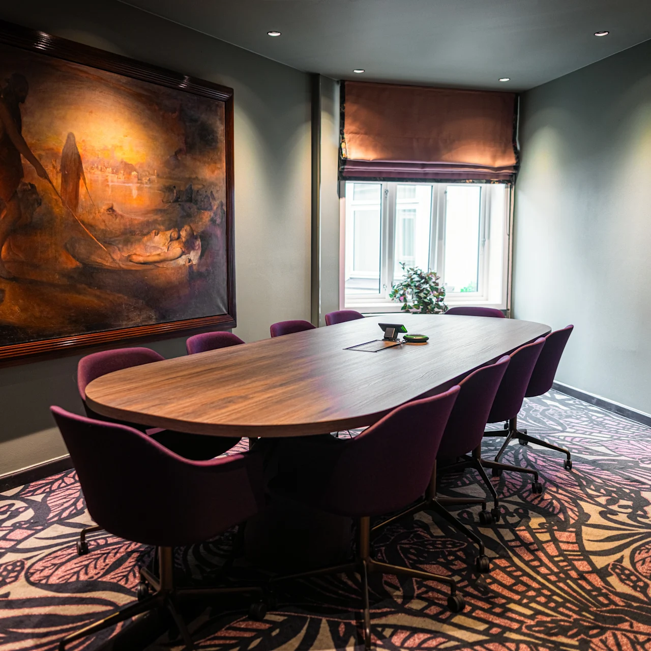 A long oval table with purple chairs arranged around it in a conference room, featuring a large painting on the wall and patterned carpet underfoot.