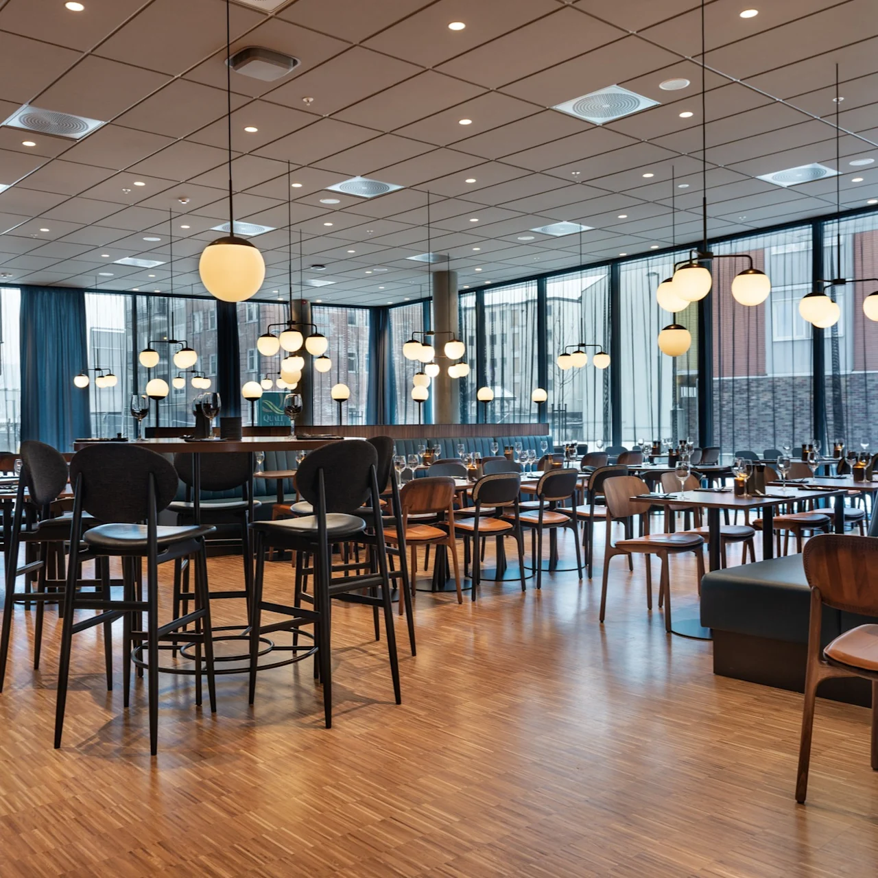 En moderne restaurant med tregulv og mørke møbler har bord dekket med vinglass. Hengende sfæriske lamper lyser opp det romslige interiøret, omgitt av store, draperte vinduer.