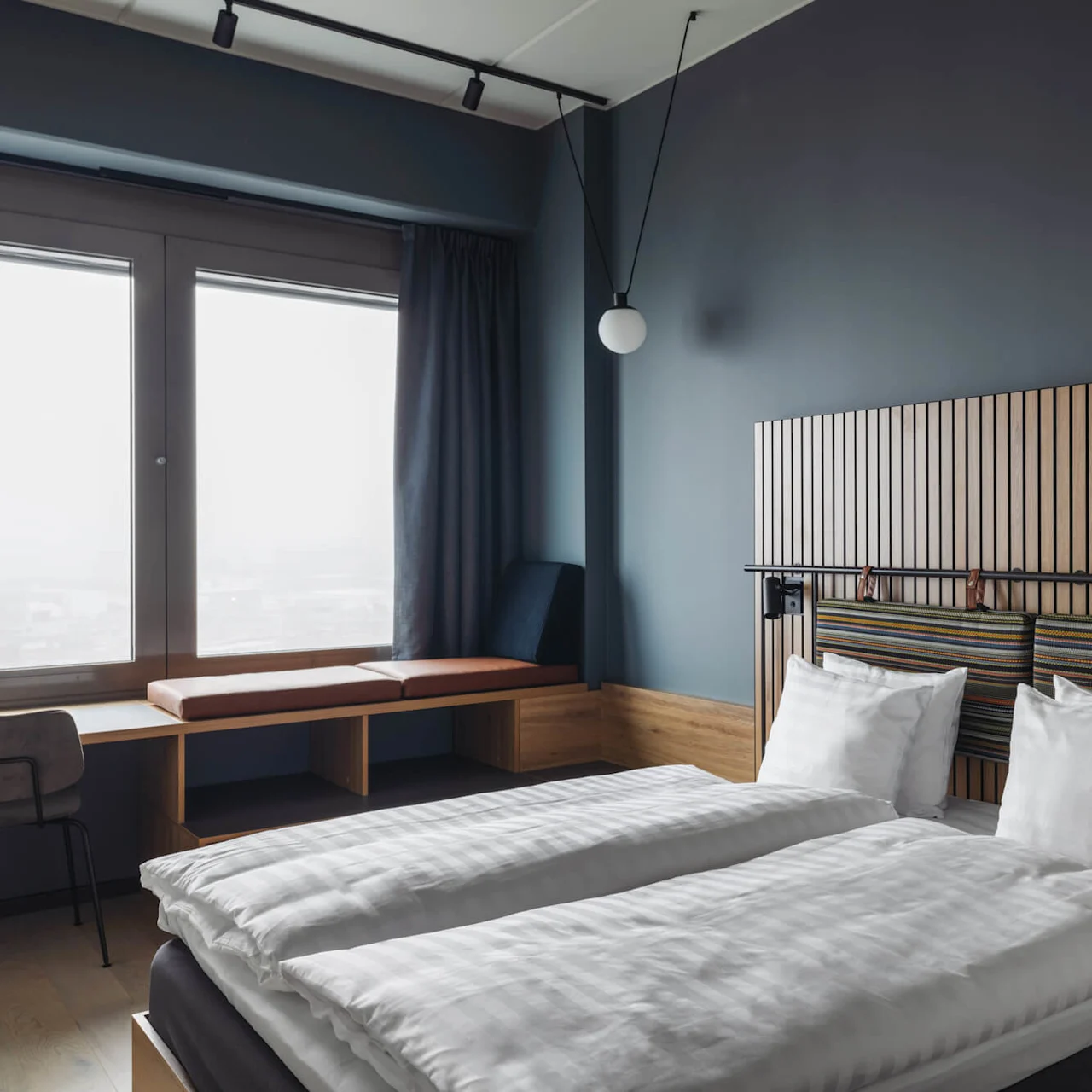 A double bed with white linens is flanked by a wooden headboard with striped cushions. Nearby, a window seat and desk sit by large windows in a modern room.