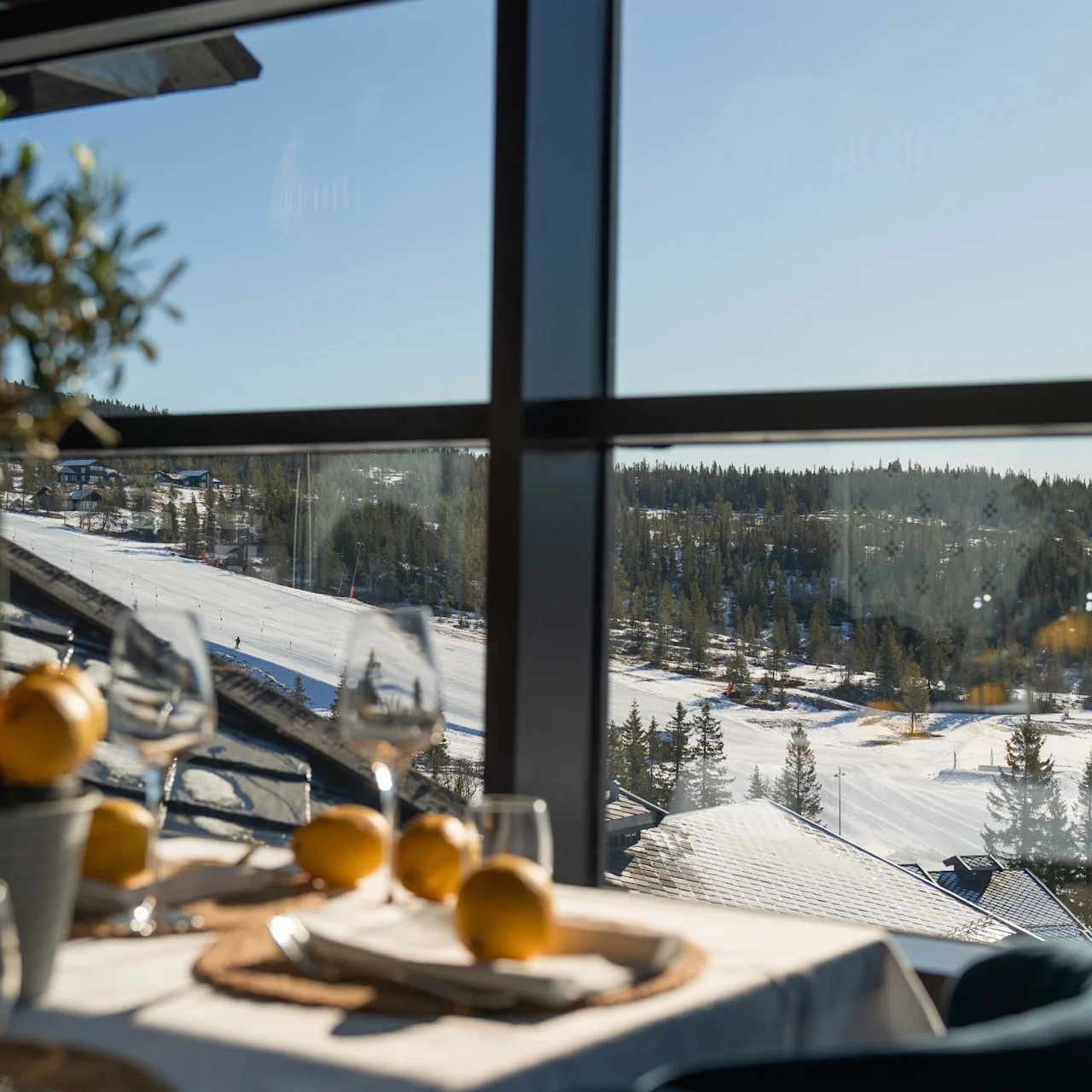 Table set with lemons and wine glasses, beside a large window. Outside, a snowy ski slope stretches across a forested landscape under a clear blue sky.