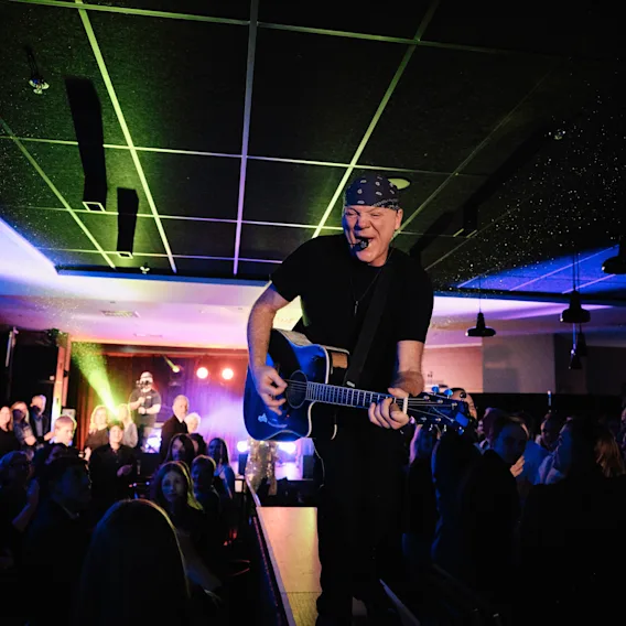 En musiker på scenen, spiller akustisk gitar og synger i en mikrofon, med fargerike scenelys og et publikum i forgrunnen.