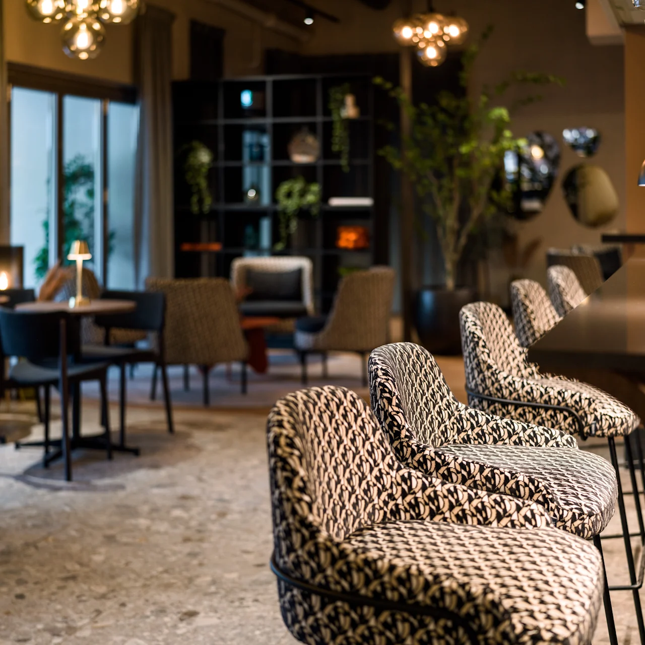 Elegant bar stools are positioned along a sleek counter in a stylish dining area; the background features tables, chairs, and decorative lighting, creating a sophisticated ambiance.