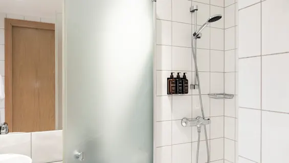 En ren, modern duschplats med vita kaklade väggar, ett duschhuvud och tre flaskor toalettartiklar. En frostad glasvägg syns.