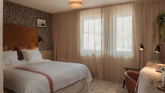 A cozy hotel room with a large bed, a desk area, and bright windows with sheer curtains, suggesting comfort and relaxation.