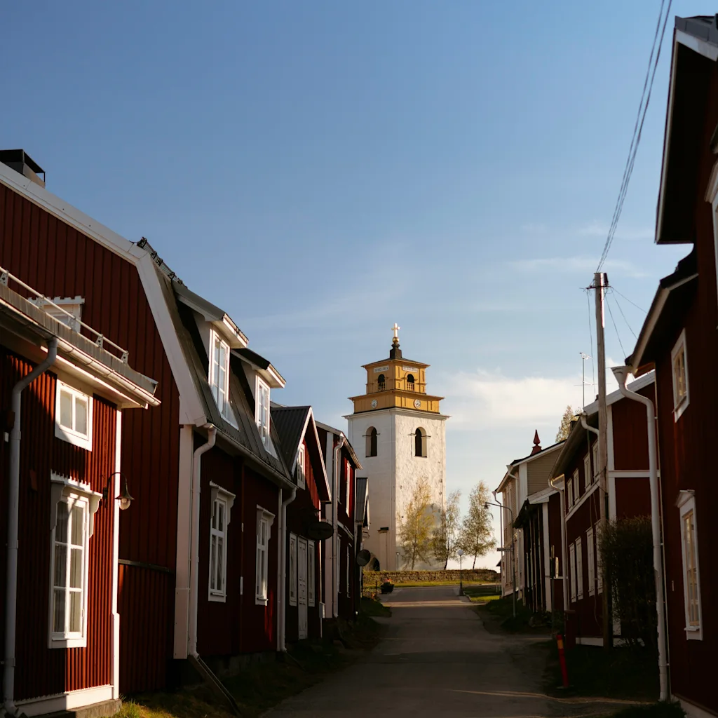 A narrow street lined with traditional red wooden houses leads to a white church tower under a clear blue sky.