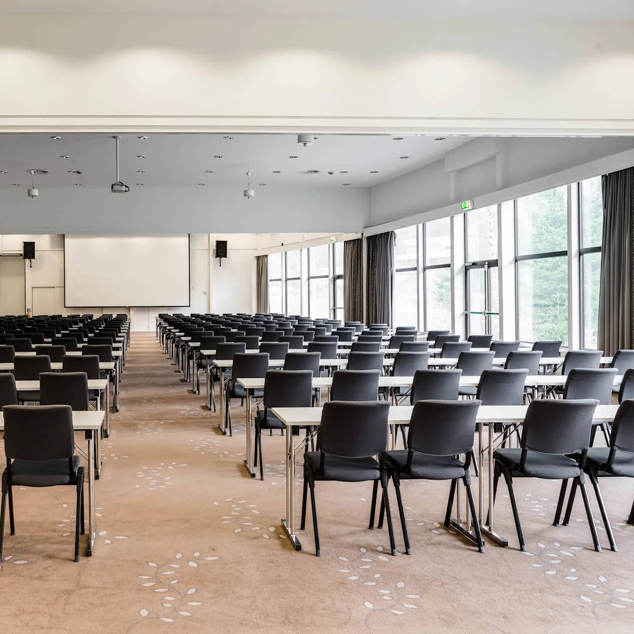 Rows of empty black chairs face a large screen in a spacious conference room with beige carpet and natural light from tall windows.