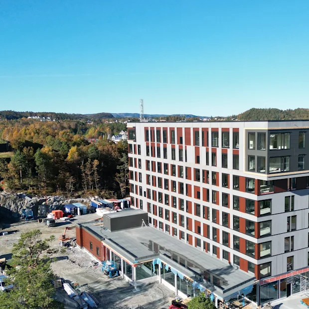 En moderne bygning under opførelse med klarblå himmel og efterårstræer i baggrunden, omgivet af byggeaktivitet.