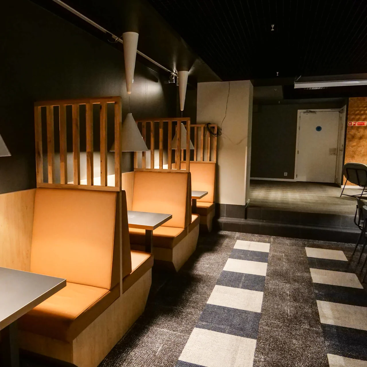 Modern booths feature orange seating and grey tables, with hanging cone lamps, beside a hallway in a dimly-lit restaurant or café. A bottle rests on the table.