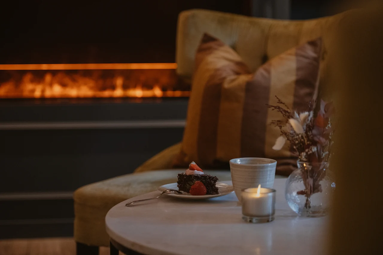 A cozy scene at Home Hotel Kung Oscar with a slice of cake, a warm drink, and a lit candle by a fireplace.
