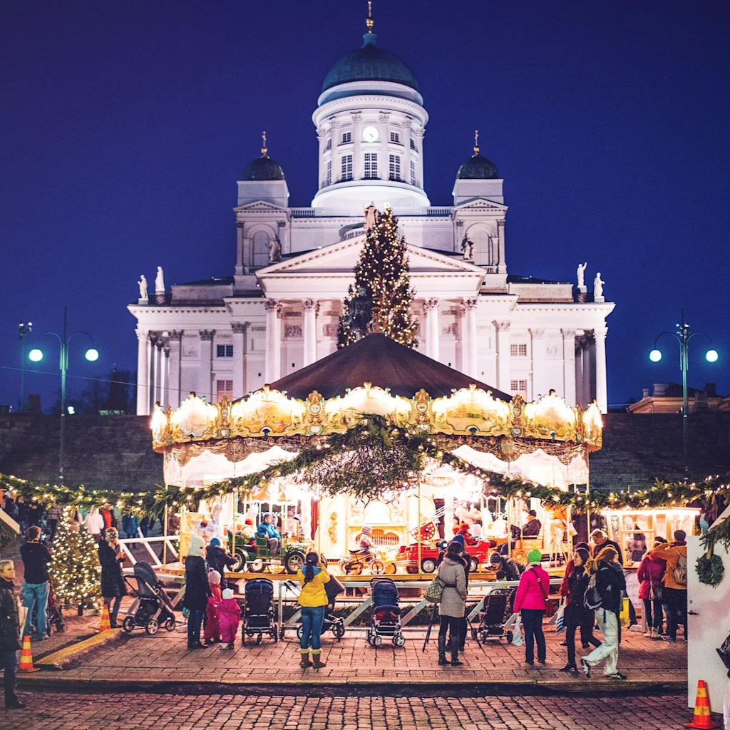 A festive Christmas market at Senate Square in Helsinki at night, with a carousel, stalls, and people enjoying the atmosphere.