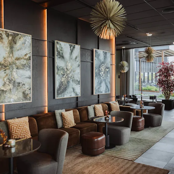 Koselig lounge med brune sofaer og mønstrede puter, abstrakt kunst på mørke vegger, og runde bord. Unike pendellamper henger over, og store vinduer viser en hage.