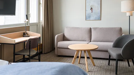 A neatly arranged room features a gray sofa facing a circular wooden table. A desk with a lamp and chair is nearby, and a window lets in natural light.