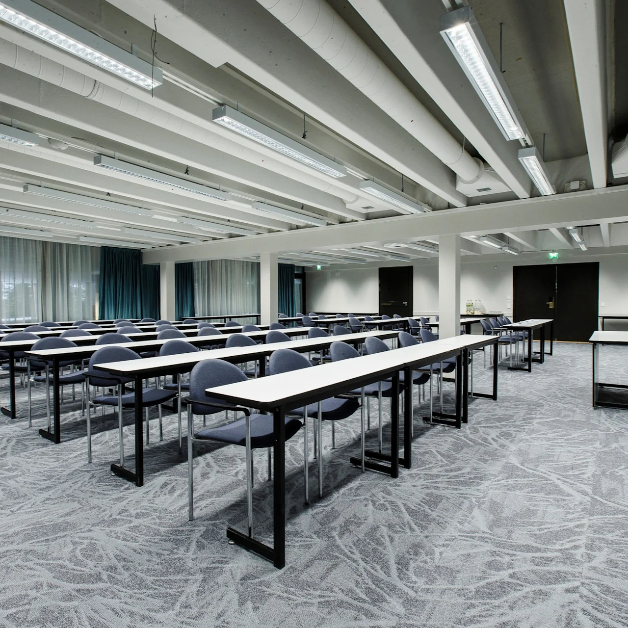 Rows of empty desks with chairs arranged in a meeting room. Bright overhead lights illuminate the space. A flip chart stands in the corner.