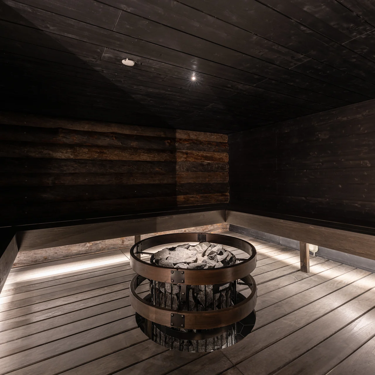 A fire pit with stones is centrally placed in a dark wooden sauna, surrounded by wooden benches. Soft lighting illuminates from beneath the benches, enhancing the cozy atmosphere.