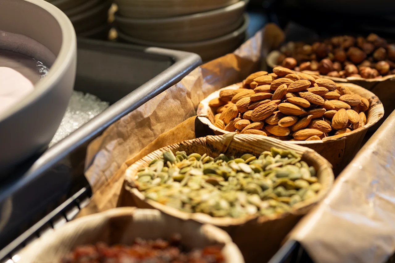 Skålar med mandlar, pumpafrön och blandade nötter står på en disk, omgivna av andra skålar, och skapar ett inbjudande urval av snacks i en matmarknadsmiljö.