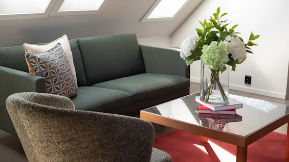 En koselig stue med en grønn sofa, en lenestol, et salongbord med blomster og bøker, og lyse takvinduer.