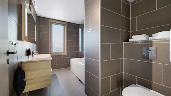 Moderne hotellbaderom med grå fliser, lyst treverk, vask og et badekar ved vinduet.