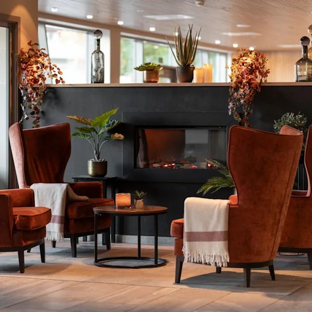 En hyggelig hotellounge med fire orange lænestole, en tændt pejs og dekorative planter, der indbyder til afslapning.