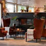 En hyggelig hotellounge med fire orange lænestole, en tændt pejs og dekorative planter, der indbyder til afslapning.