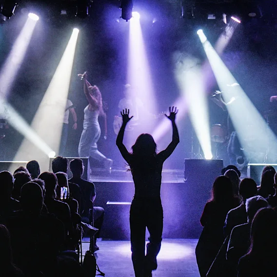 En livlig konsertscene med publikum som ser artister på scenen under lilla og hvite scenelys. En person i publikum har hendene hevet.