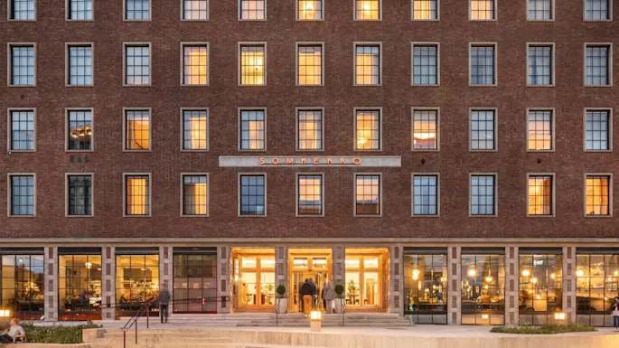 A brick building with lit windows, people entering its brightly illuminated entrance. The sign above reads "SOMMERRO." Surrounding area features steps and landscaped bushes.