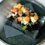 En ret viser delikat anrettet røget laks toppet med creme, gule bær og mikrogrønt, placeret på en glasblok og serveret på en sort tallerken.