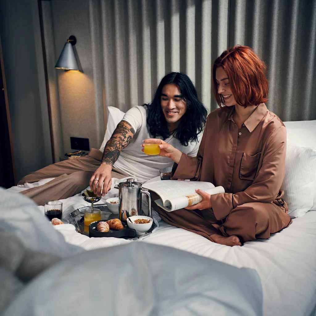 Två personer njuter av frukost på sängen, en häller upp juice och den andra läser en bok, med croissanter och kaffe på en bricka.