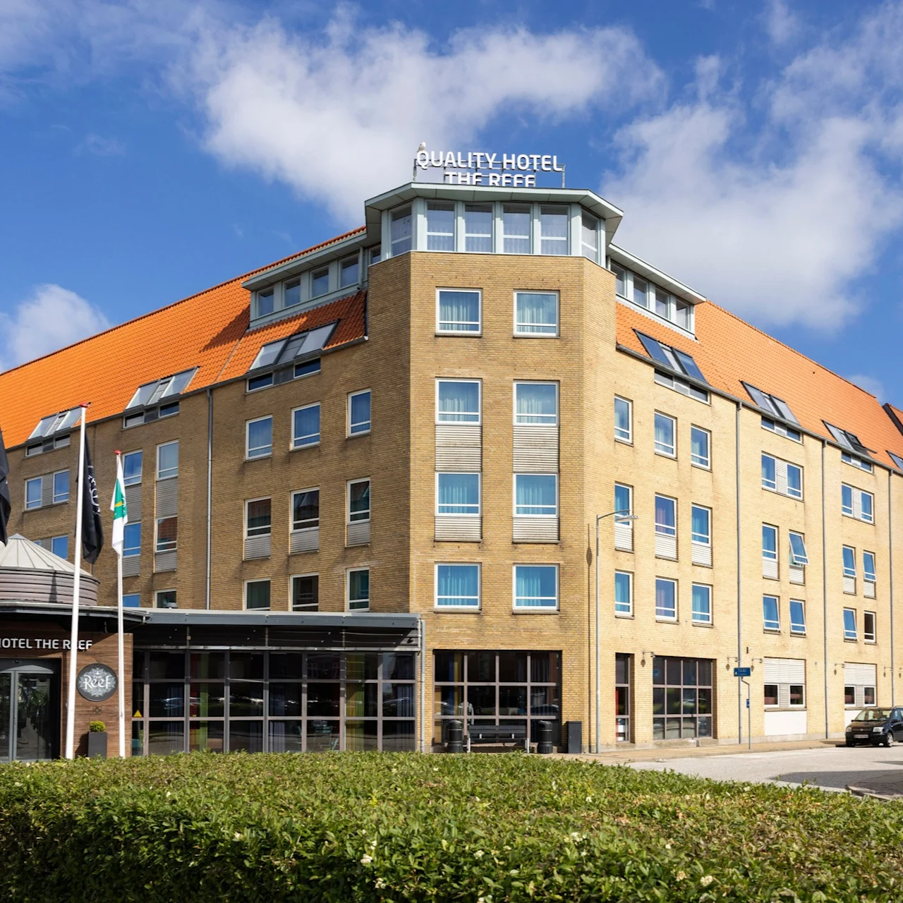 A large brick building with a red-orange roof labeled "Quality Hotel The Reef" has several windows and flags in front, situated on a bright day with a partly cloudy sky.