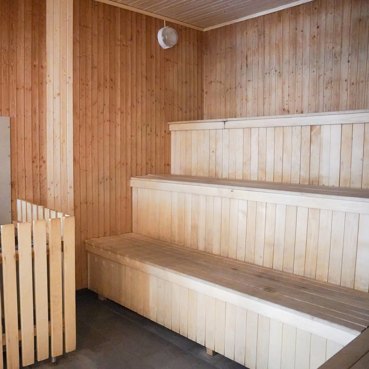 Wooden benches sit empty in a sauna room with paneled walls, a heater unit enclosed by a wooden fence is visible nearby.