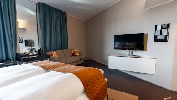 Bed with white linens and orange cover, facing a wall-mounted TV; next to it, a sofa near a long curtain. Wall-mounted cabinet beneath TV. Room has neutral decor.