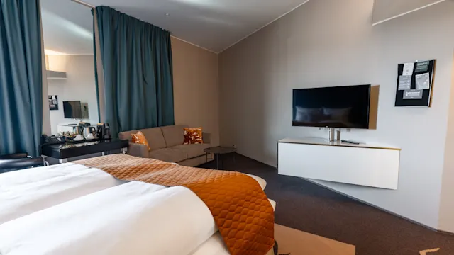 Sänky valkoisine vuodevaatteineen ja oranssi päiväpeite, jonka edessä seinään kiinnitetty TV; vieressä sohva pitkän verhon lähellä. Seinään kiinnitetty kaappi TV:n alla. Huone on neutraalin sisustuksen.