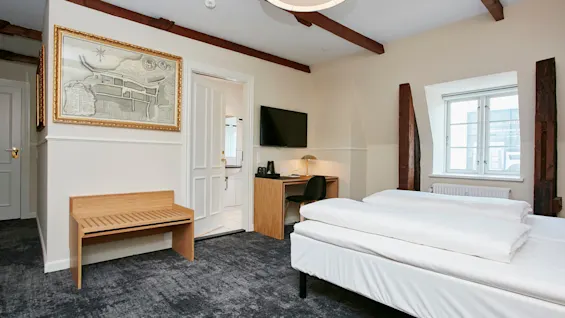 Et hyggeligt hotelværelse med en komfortabel seng, et lille skrivebordsområde, et vægmonteret tv og rustikke synlige loftsbjælker.